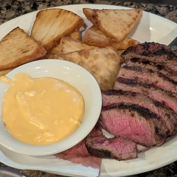 Sliced steak with a pink center, a side of fried potatoes, and a bowl of cheese sauce.