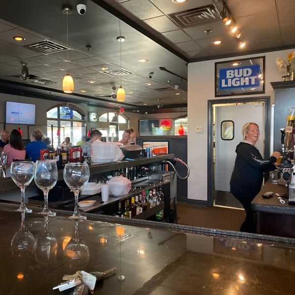 A bartender smiles while working behind the bar at Reifschneiders Grill and Grape, with patrons in the background.